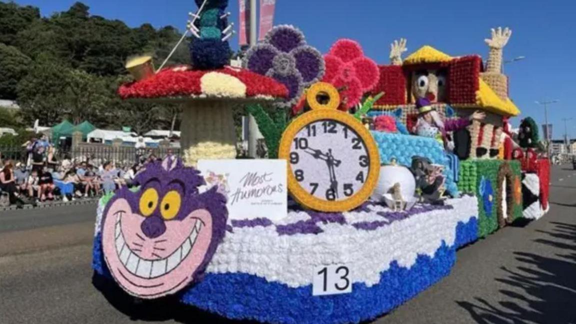 A colourful Alice in Wonderland themed float is on the road with a crowd watching to the left. The float is adorned with colourful flowers depicting a Cheshire cat decorated with a clock, mushrooms, flowers and an oversized Alice in a small house. 