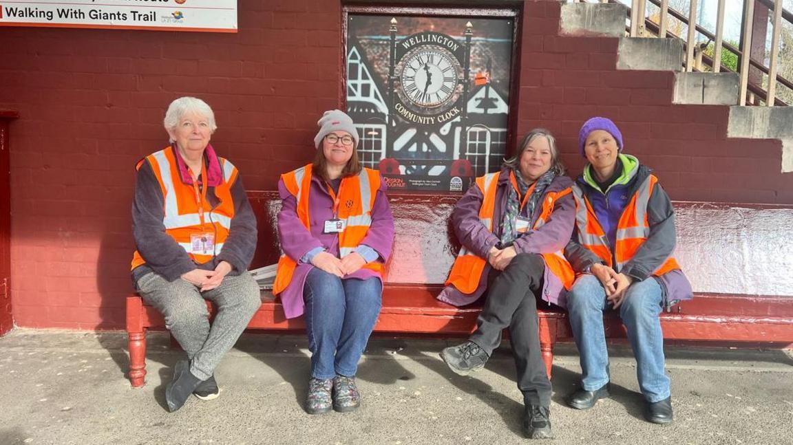 Four women in orange reflective jackets sat on a red bench in front of a red wall with a set of steps running up one side. There is an image of a clock set in front of a black and white timbered building on the wall behind them.