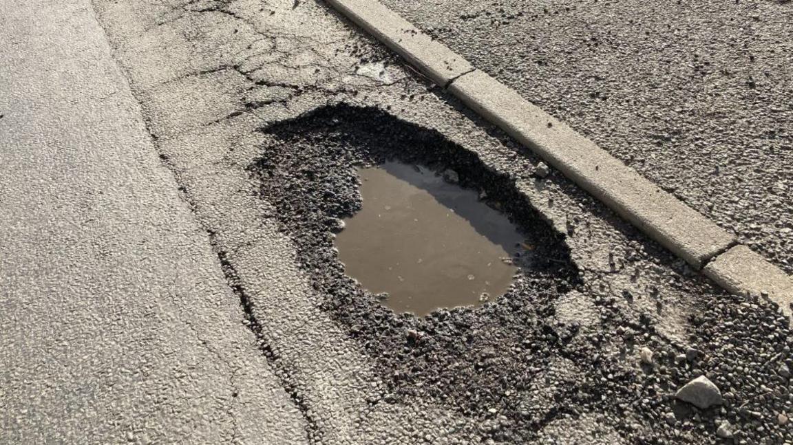 A deep wide pothole in the road next to a curb full of water, surrounded by cracked tarmac