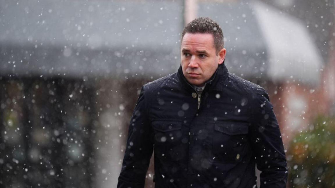 A man with short dark hair and wearing a dark jacket walks through falling snow.