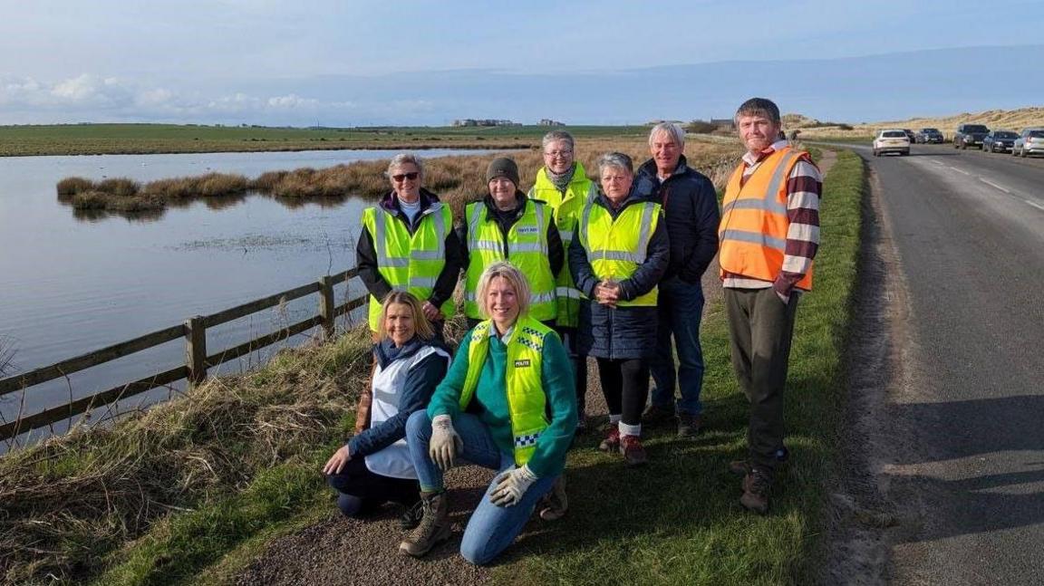 A group of eight people, two kneeling, are gathered by a wooden fence with a pond to the left and a road to the right. They are wearing hi-vis vests. In the distance Bamburgh Castle can be seen on the horizon.