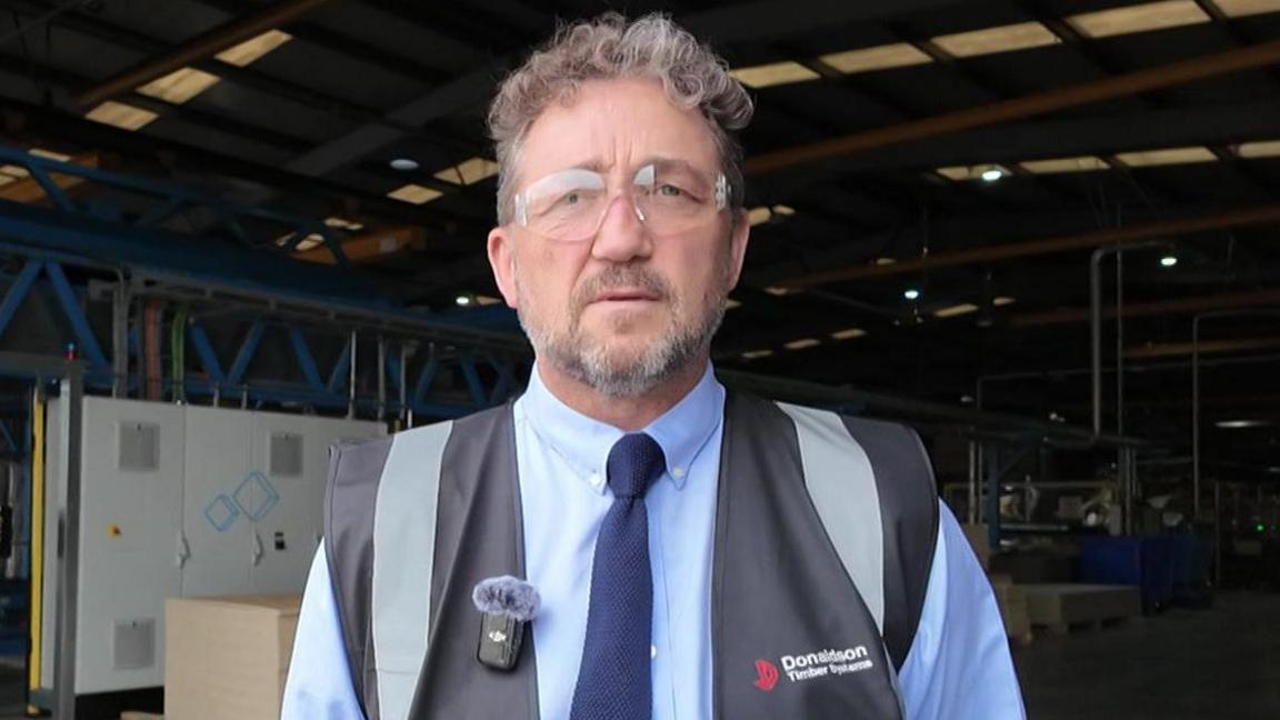 Alex Goodfellow has short brown hair and grey stubble, and is standing outside of a factory. He is wearing a grey hi-vis jacket over a blue shit and navy blue tie.