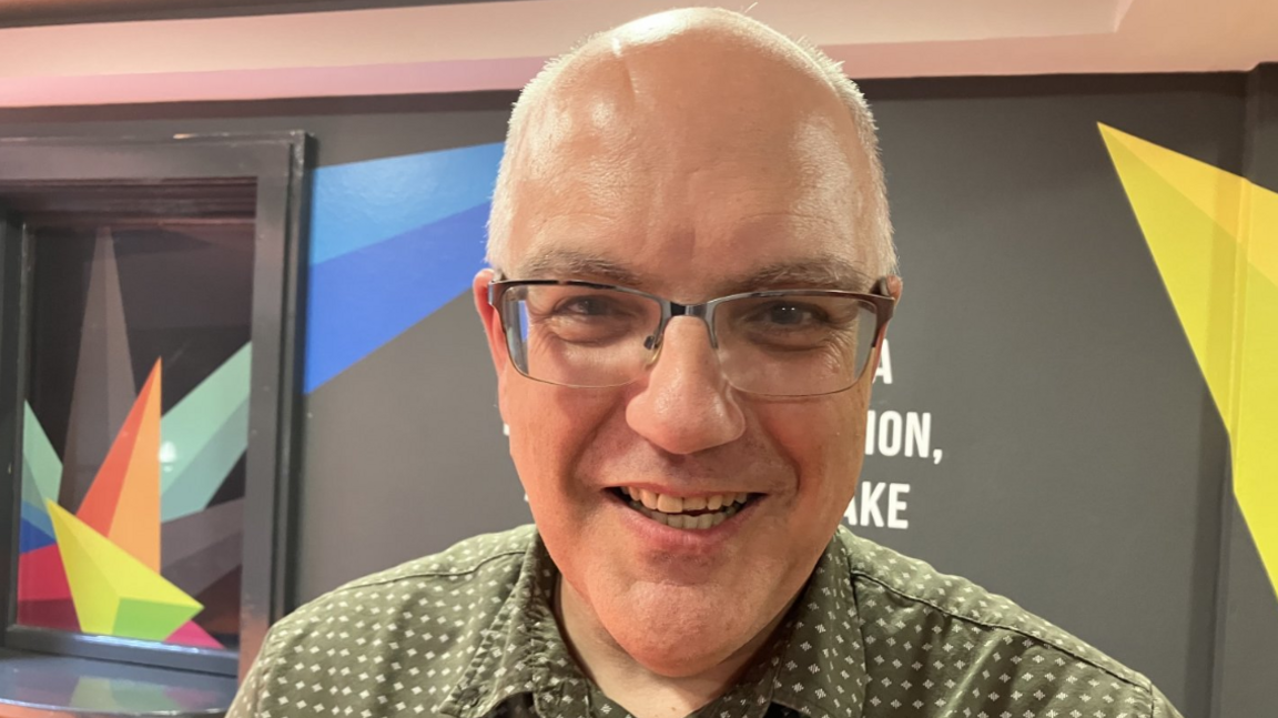 Shows a man with a bald head and glasses smiling. He is wearing an olive green open neck shirt which is covered in little stars.
