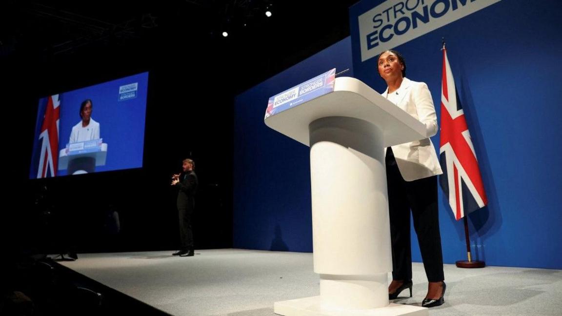 Kemi Badenoch in white jacket and black trousers and heels at a podium in front of a union flag