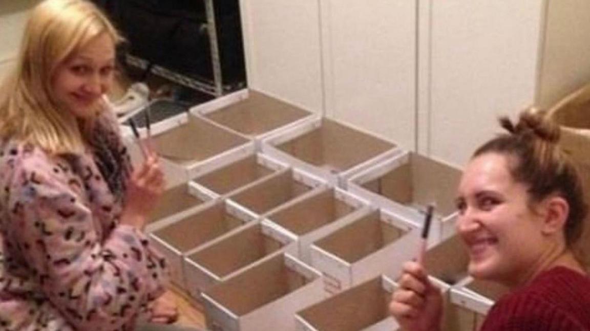 Sophie and Hannah in a garage with boxes. The are looking at the camera and smiling and holding make-up brushes,