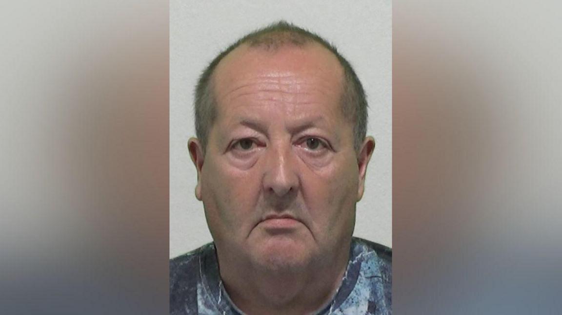 Mugshot of John Briggs. He has very short balding dark hair, is clean shaven with a large head and downturned mouth. He is looking very morose.