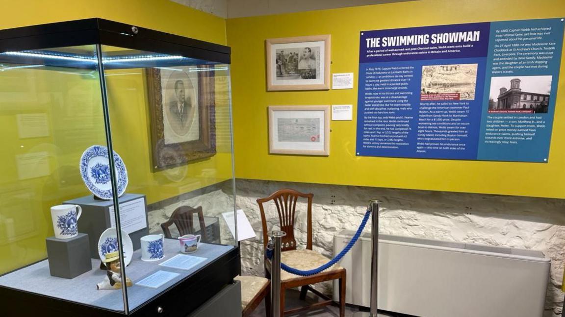 A wide shot of the corner of an exhibition, on the left of the picture is a see-through display case with plates and cups inside. Next to it on the wall there are some framed pictures of Captain Matthew Webb. On the right of the picture there is a blue display board bearing the words The Swimming Showman with two black and white pictures and three columns of writing in a white font which is not legible .