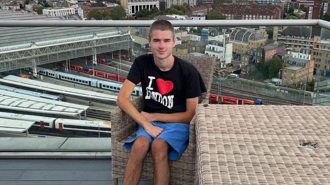 Liam Virgo wearing a black t-shirt and blue shorts sitting on outdoor furniture on a terrace that overlooks St Pancras railway station in London