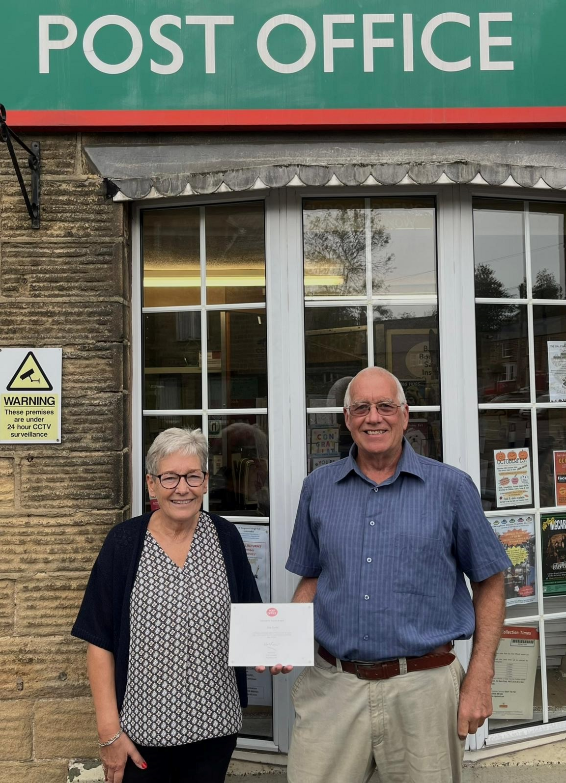 Mrs Woodward (left) and Mr Dolby stand smiling outside the front of their post office, with a large window behind them. They are holding a long service certificate from the Post Office. Mrs Woodward has short grey hair and wears glasses. She is wearing a dark cardigan, dark trousers and a patterned top. Mr Dolby wears glasses, has short grey hair and is wearing a blue shirt with light colour trousers