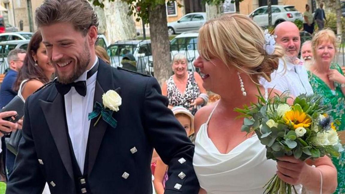 Callum Kerr, a dark haired man wearing a kilt, smart jacket and a bow tie with a white rose as a button hole. He is holding the hand of an older woman, his mother, who has blonde hair and is wearing a wedding dress. She is holding a bouquet of flowers. Both are smiling.