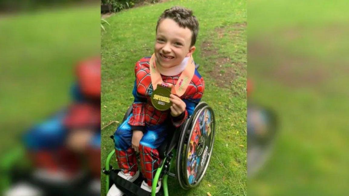 A young white boy with dark hair sits in a bright green wheelchair dressed as Spiderman in a blue and red suit. He smiles and holds up a large gold medal.