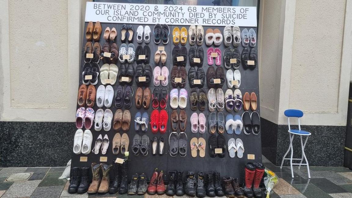 The display, which shows a variety of shoes, including flip flops, trainers, boots and high heels, on an upright board. Each has the names of different people, the sign reads between 2020 and 2024 68 members of our island community died by suicide confirmed by coroner records.