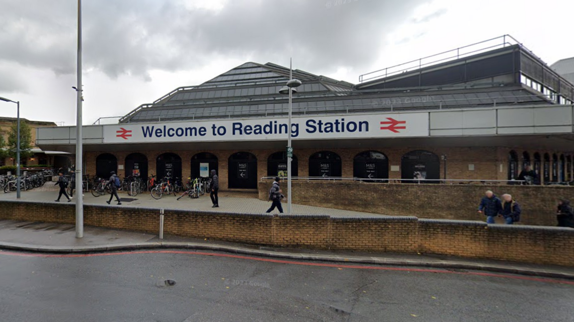 The front of Reading railway station, a brick building with a large sign outside reading "welcome to Reading Station". There are people walking outside and bikes locked up by the entrance.