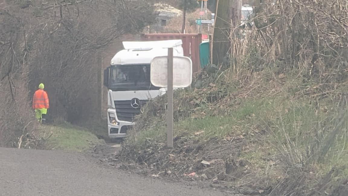 A lorry stuck on a small road with a person in high-vis clothing nearby. 