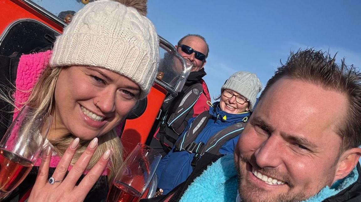 Ms Francis and Mr Tite are both smiling into the camera after the proposal. Ms Francis is wearing a pink and black Dryrobe and is showing her wedding ring off. She is holding a glass of champagne. She has a cream bobble hat and a nose ring. Mr Tite is wearing a blue and black Dryrobe and is holding a glass of champagne. 