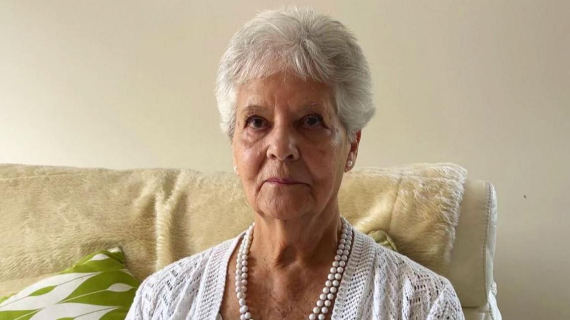A recent picture of Ann, she has short grey hear and is wearing a white cardigan and a pearl necklace.