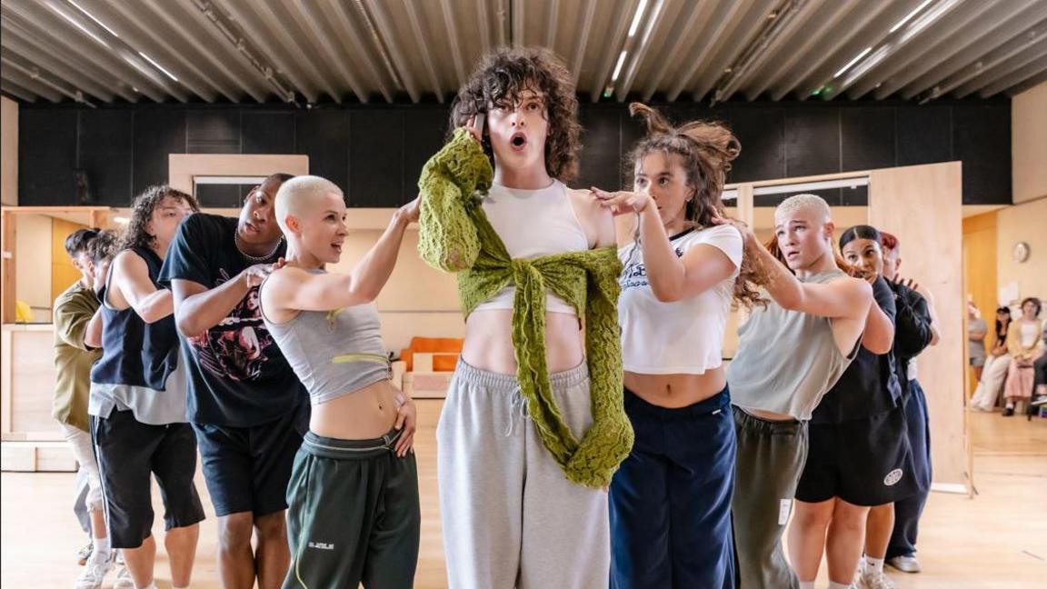 An actor holds a phone up to their ear while wearing a white tank top and green scarf. They have curly hair. Behind them are six back up dancers in baggy t-shirts and sportswear.