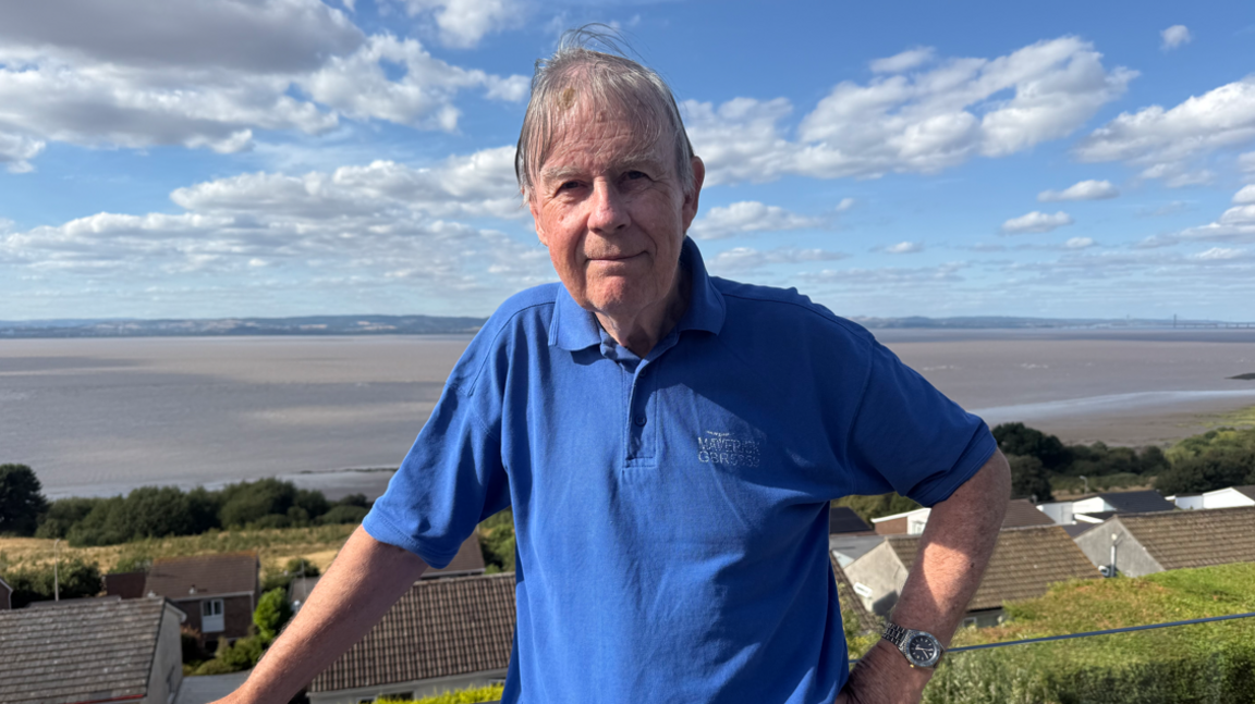 Huw looks at the camera, with the sea behind him. He has short grey hair and wears a blue polo shirt.