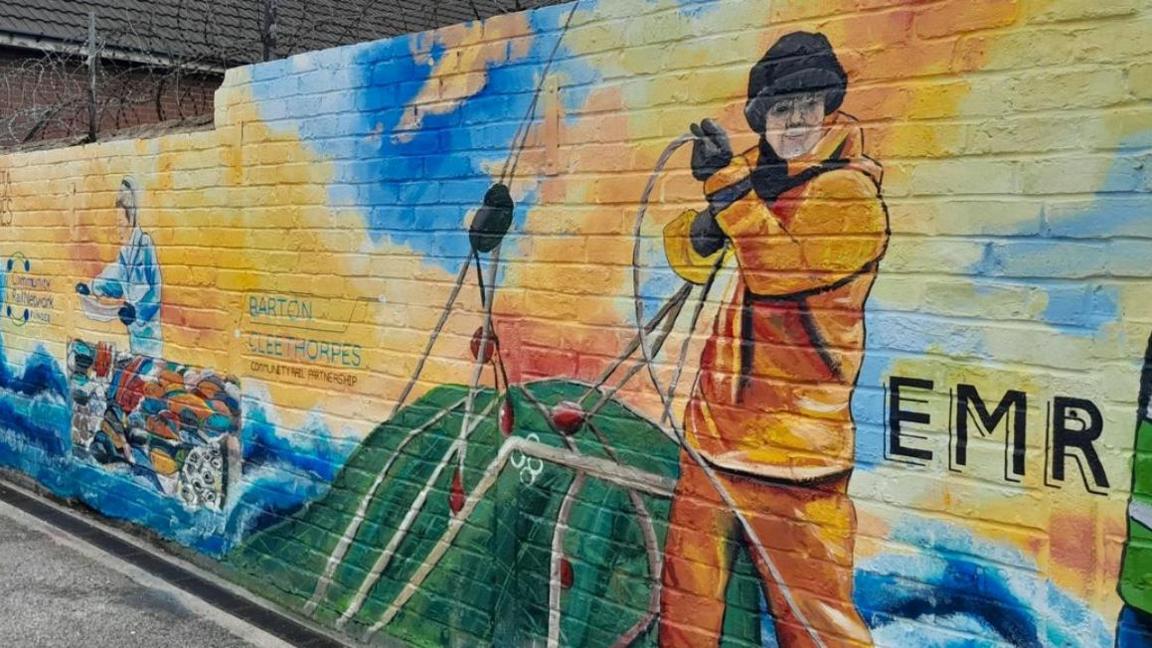 Mural depicting a fisherman hauling in nets with another figure in the background sorting the catch. The letters EMR are painted in black on the right edge of the artwork.