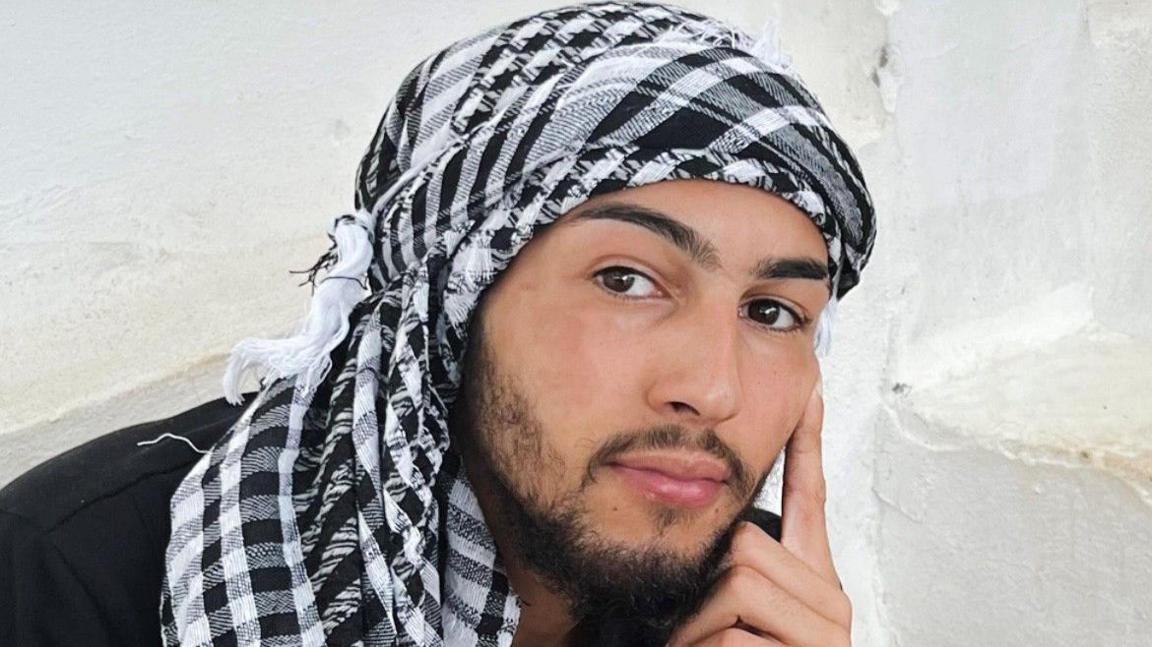 Marley Gascoine wearing a black and white head scarf Keffiyeh. He has a short dark beard as well.