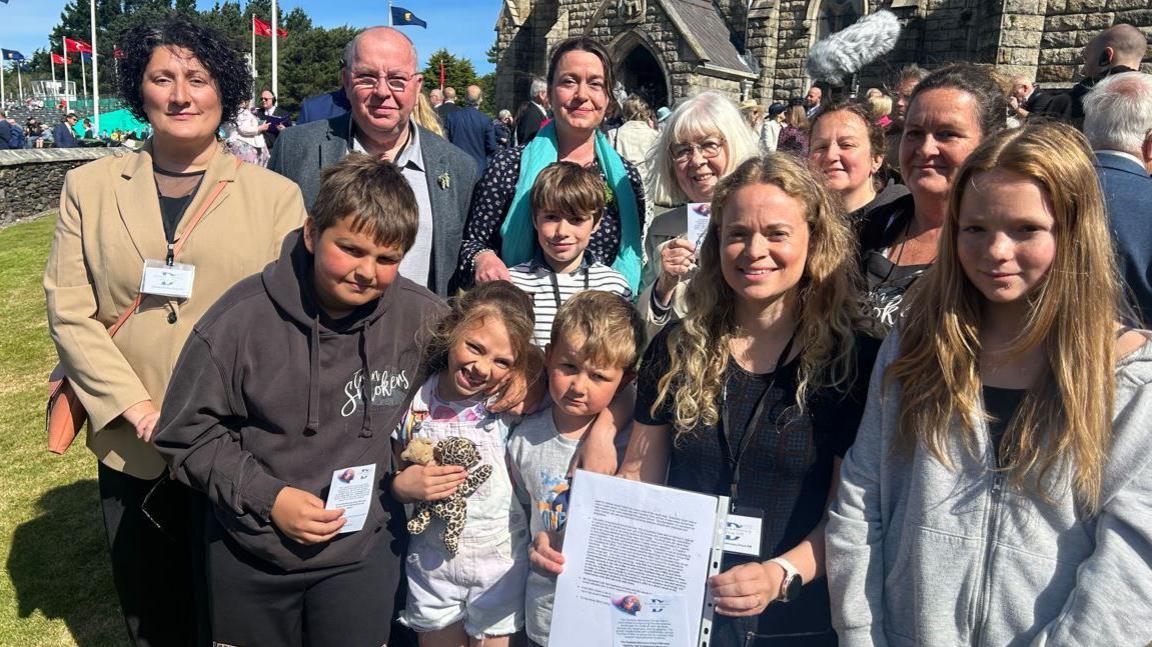 A group of petitioners, children and adults, gather together. Julia Bell holds the petition to call for more school support for children with dyslexia which is printed on white paper and is in a plastic folder.