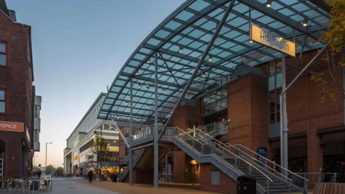 Exterior shot of Harlequin Theatre showing large glass covering and metal staircase leading up to entrance