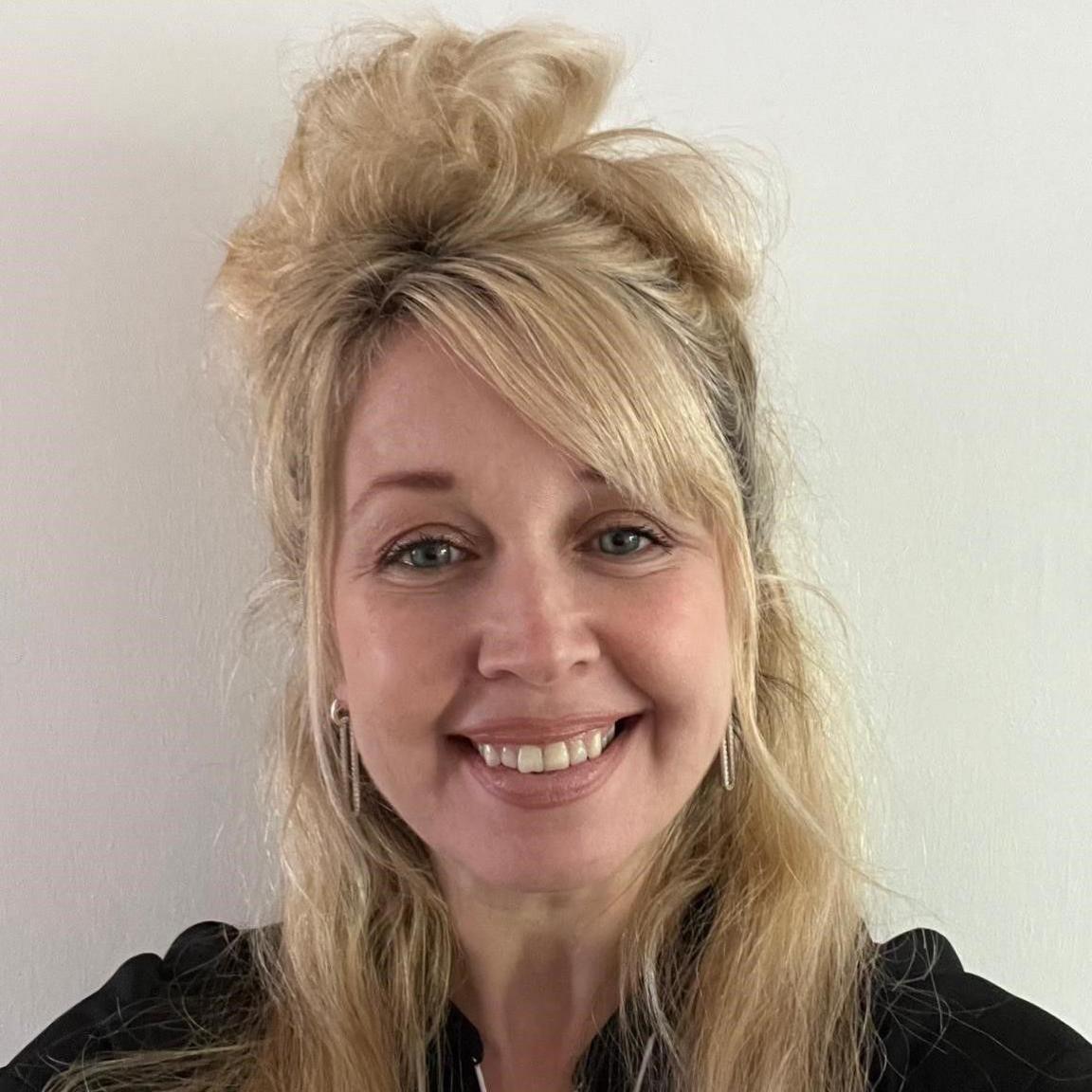 A woman with long blond hair, standing in front of a cream background, smiles at the camera