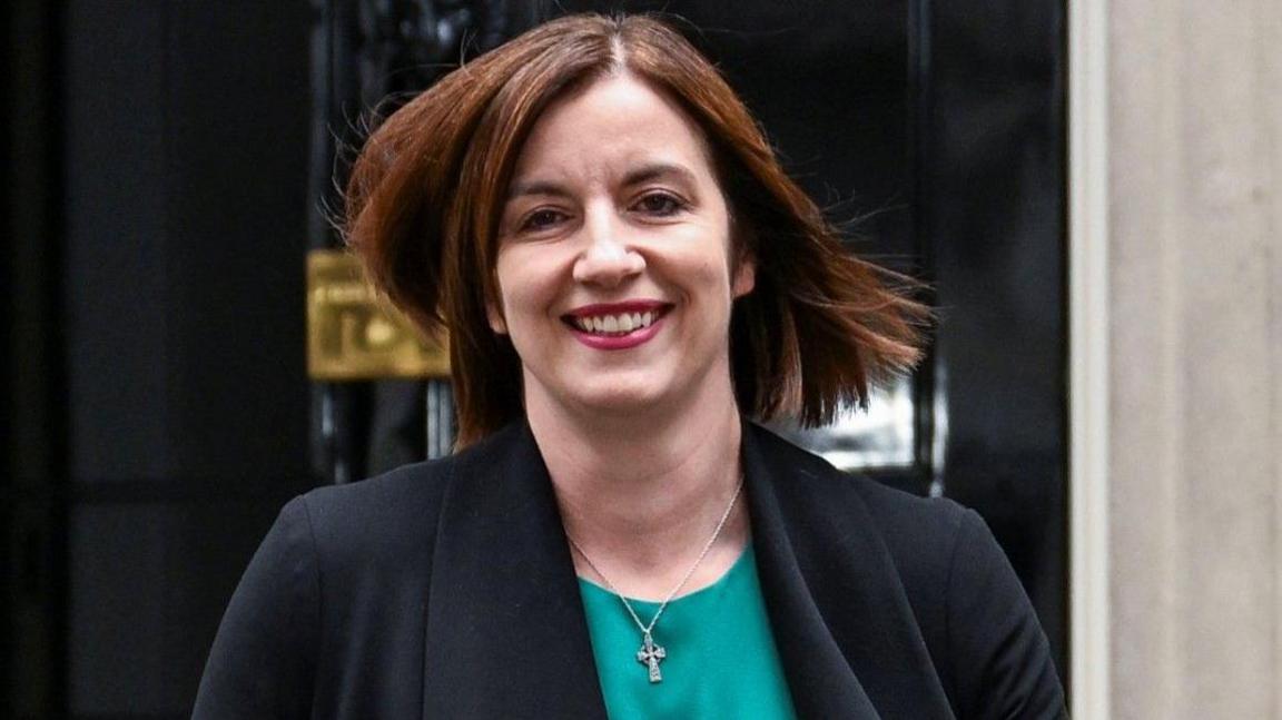 Phillipson wears a teal scoop-necked top with a black jacket and a small silver Celtic cross. She wears dark pink lipstick and is smiling. Her light brown hair is worn loose, straight and shoulder-length.