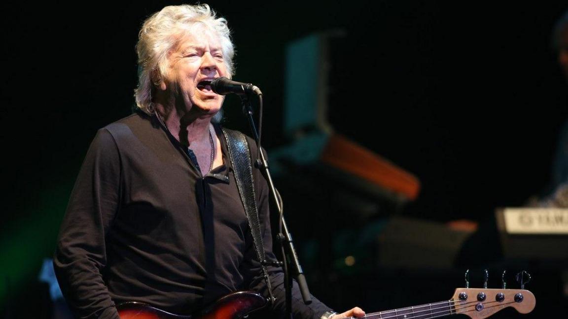 A man with white hair is singing into a microphone and holding a guitar.
