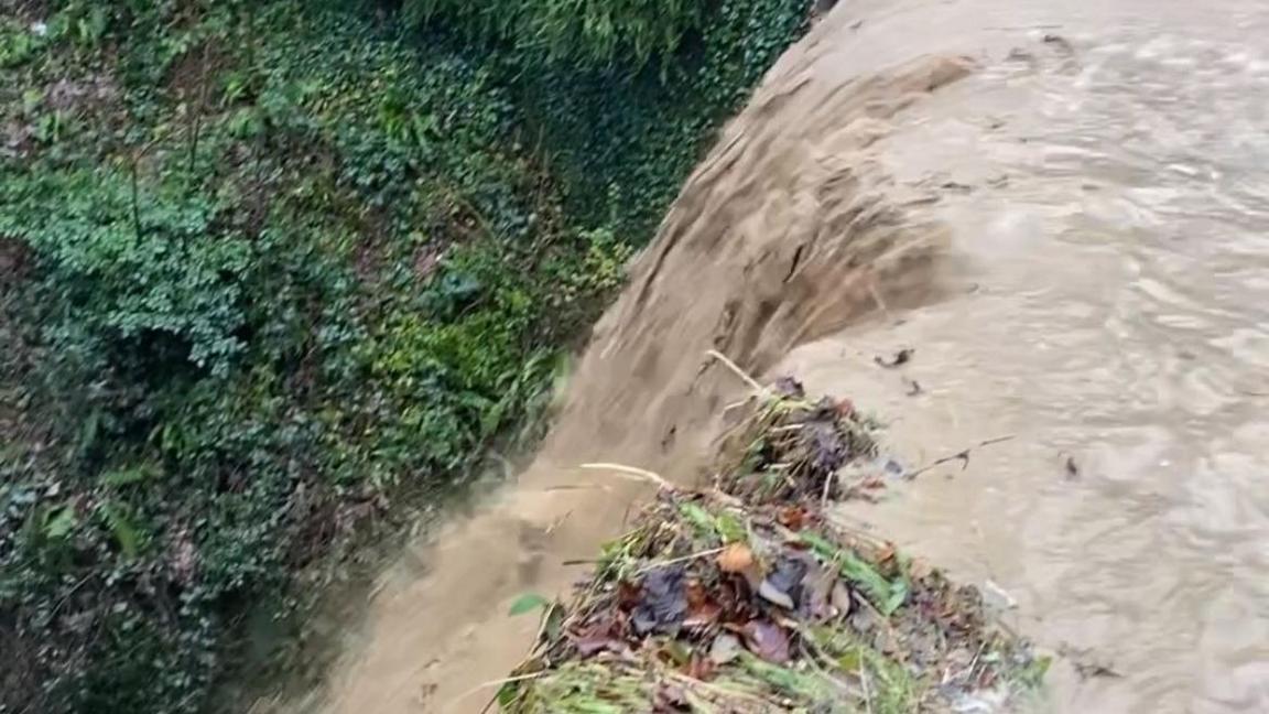 Large amounts of floodwater are seen pouring over the road in Bronygarth and over the side of the bridge. The road and bridge are not visible due to the water but some green shrubbery can be seen on the other side. 