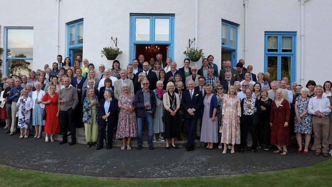 A group of smartly dressed people standing in a bunch outside Government House for a photograph. The walls of the building are white and the door and window frames are painted light blue.