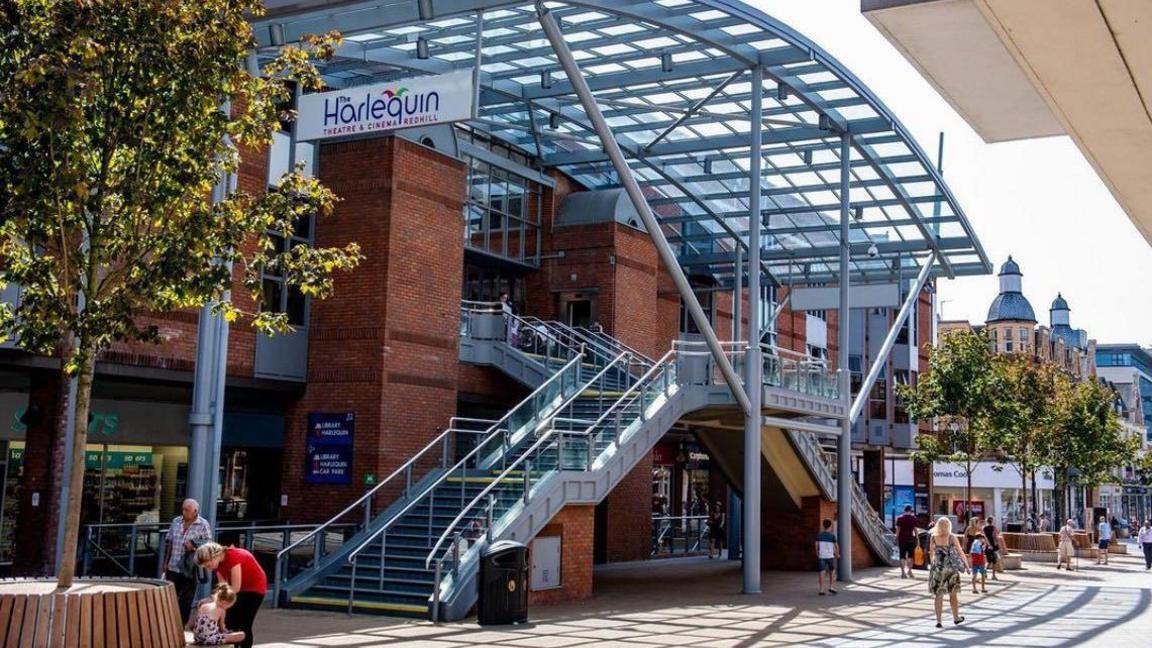 Redhill's Harlequin theatre seen from the outside. It is a red brick building with staircases running up to the doors on either side and a metal and glass canopy above it. There are people walking down the high street in front of the building, with the canopy casting a shadow on the floor in front.