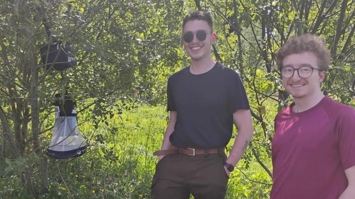 Aidan Desjardins is in a pink T-shirt and has curly brown hair and glasses.
Ryan Carmichael is wearing sunglasses and is in a black T-shirt and tan trousers with brown belt. They are both standing in a field beside a mosquito trap.