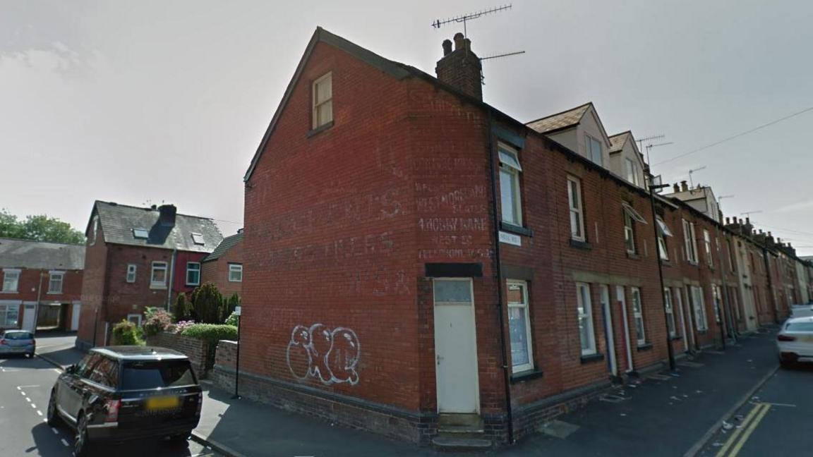 Google Streetview image of a road of red-brick terraced houses near Ecclesall Road in Sheffield, with parked cars outside the house and double yellow lines to the right. The front door is up two steps. There is white graffiti and a window on the wall on one side of the house, and two windows on the other wall with the Neill Road street sign.A chimney and aerial is on the roof.