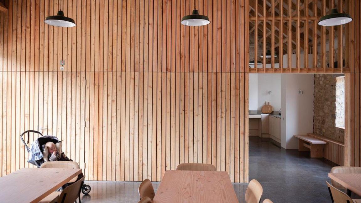 A timber panelled room with three wooden dining tables surrounded by chairs. An opening in the wall leads to a kitchen area. In the corner of the room is a baby in a push chair