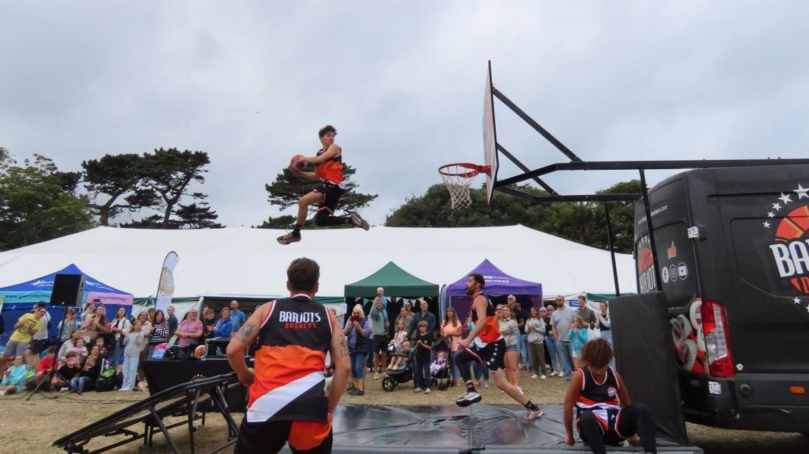 A basketball player is mid-air spinning towards a basket while attempting to do an acrobatic slam dunk. A crowd is watching in the background.