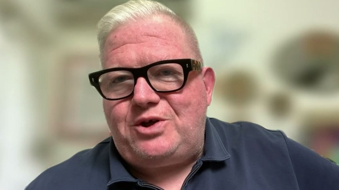 Paul Askew, who is wearing a grey-blue zip-up sweater and has short blond hair and is wearing glasses, is talking into the camera on a Zoom call.
