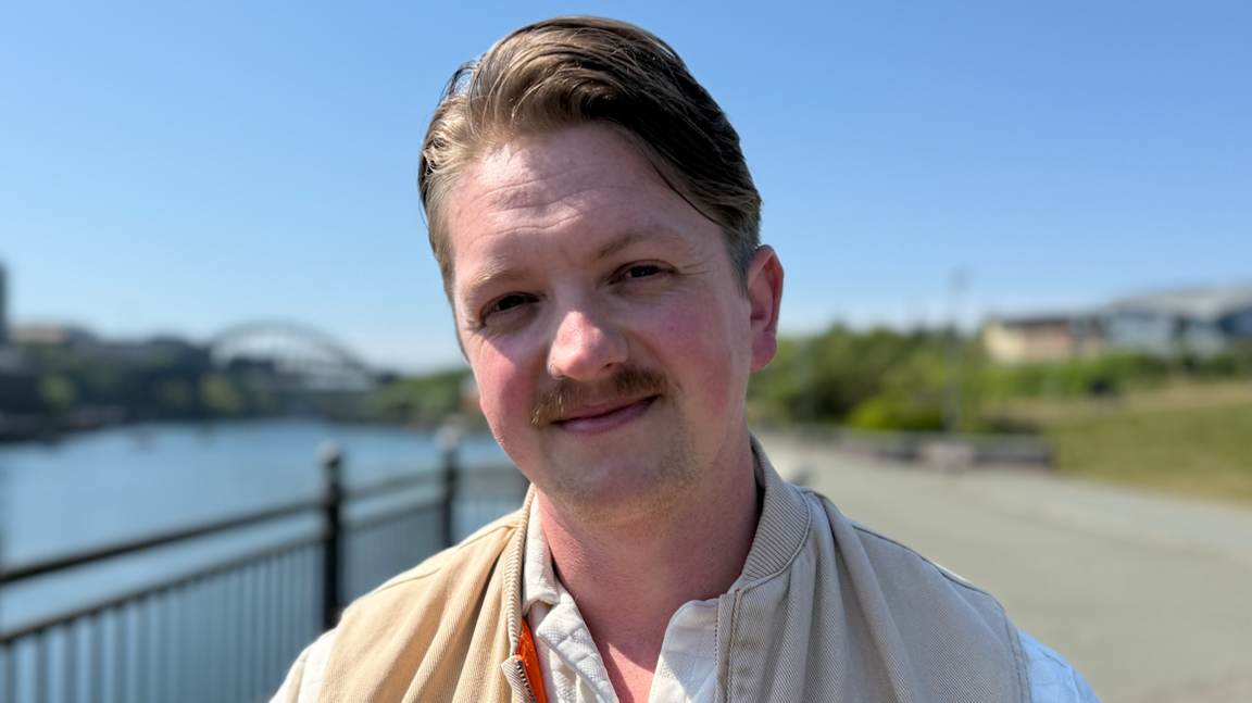 John Lee Taggart wearing a white buttoned shirt and a beige gilet style jacket stands on the Sunderland Riverside with the city's various bridges in the background. It is a sunny day.