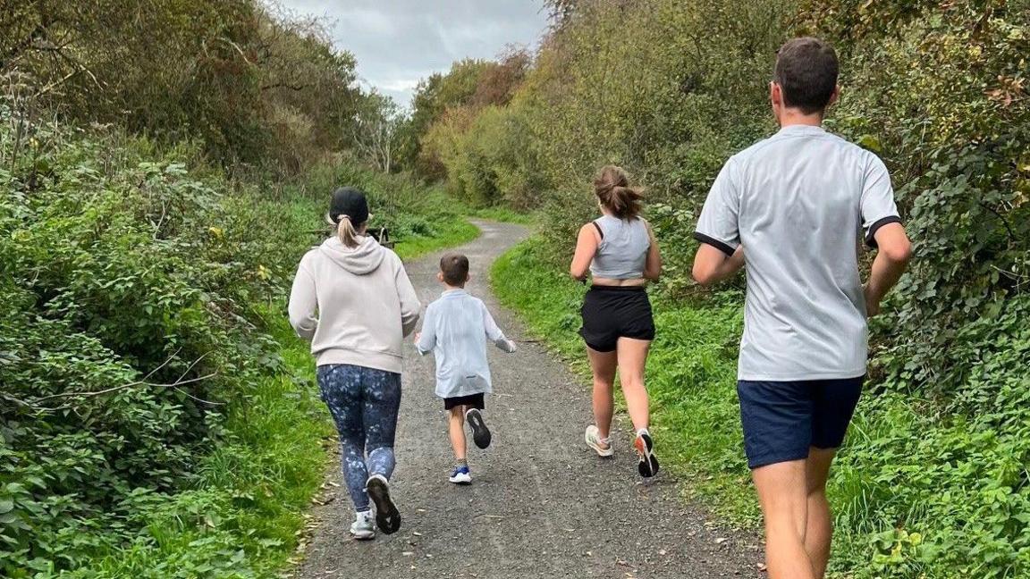 Four people are jogging along a wide dirt path surrounded by dense greenery on both sides. The path curves slightly ahead and is lined with trees and bushes. The sky is overcast with grey clouds. The runners are dressed in casual athletic wear: one in a hoodie and patterned leggings, another in a light top and shorts, and two others in shorts and T-shirts. They are moving away from the camera toward the bend in the trail.