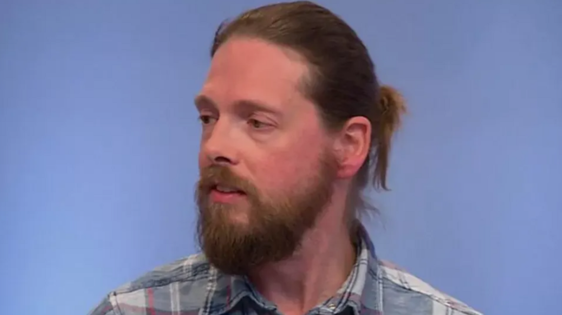 Man with a beard sitting in front of a blue background.