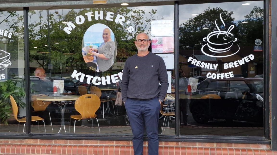 Maz Salih standing outside Mother's Kitchen, which has a glass exterior. He is wearing navy trousers, a navy jumper and glasses. 