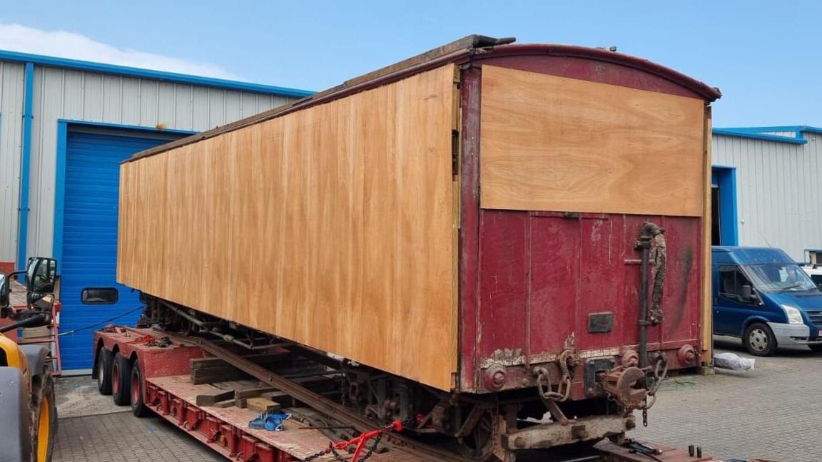 The boarded up carriage sits on the back of a truck in a car park in front of a large blue and grey storage unit.