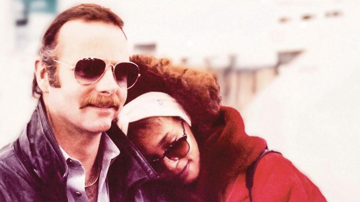 David Roberts with Whitney Houston. David has brown hair and a moustache and is wearing aviator sunglasses. Whitney has her head on David's shoulder. She is wearing dark glasses, a wide white head band and a red coat with a read fur collar.