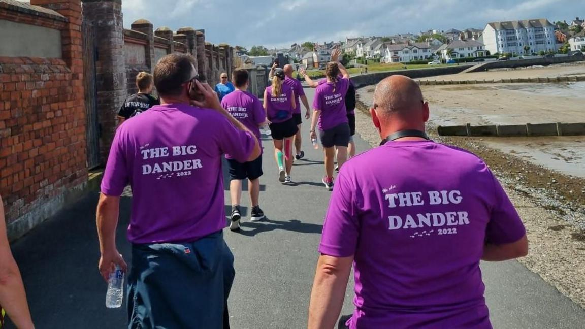 A group of people in purple T-shirts with white lettering on the backs reading "The Big Dander". They are walking along a seaside promenade, a beach is to the right and a red brick wall to the left. The sky is blue with a few clouds.