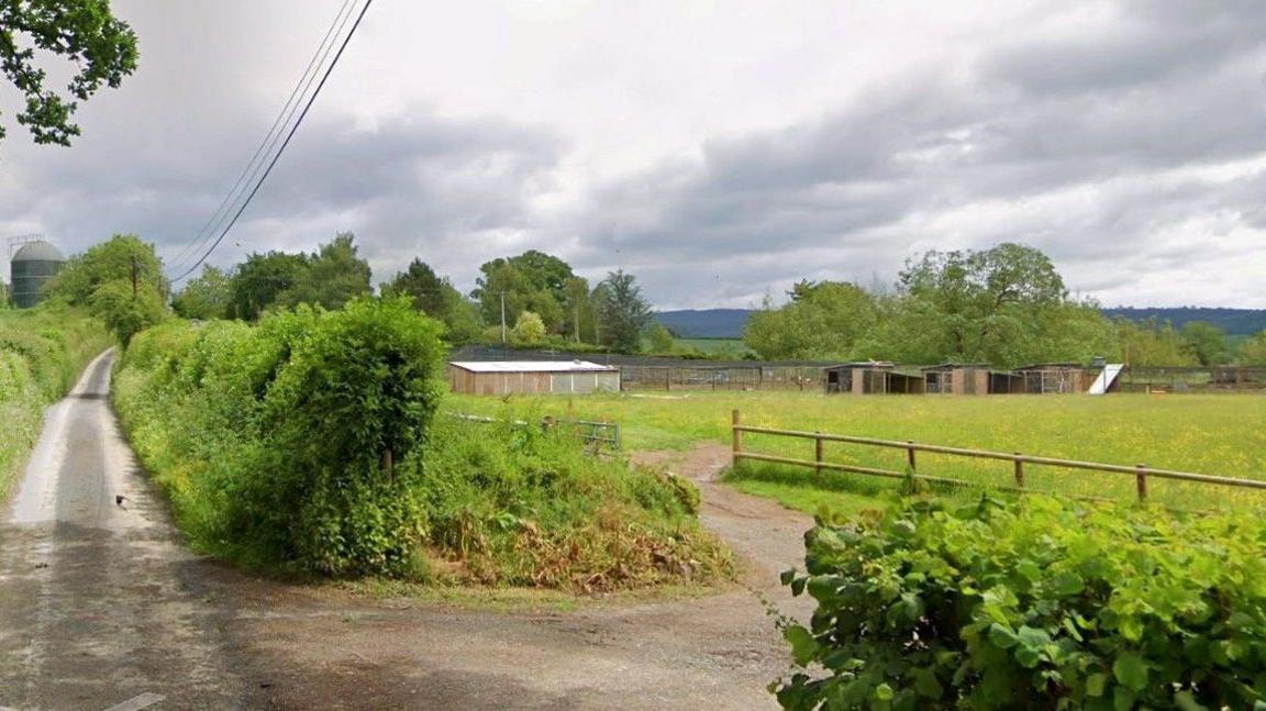 The entrance to a farm off a country lane, with hedges and wooden fencing. A field and farm buildings can be seen in the distance.