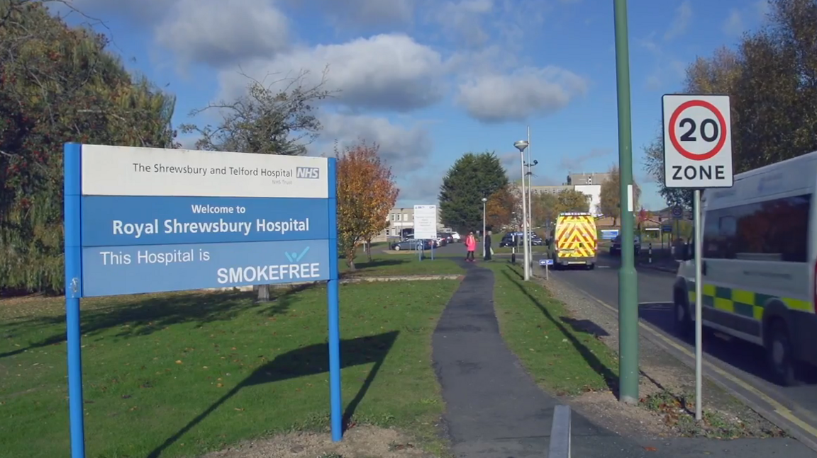 A blue and white sign for the Royal Shrewsbury Hospital. To the right of it is a road, with two ambulances driving along it, towards the hospital building 