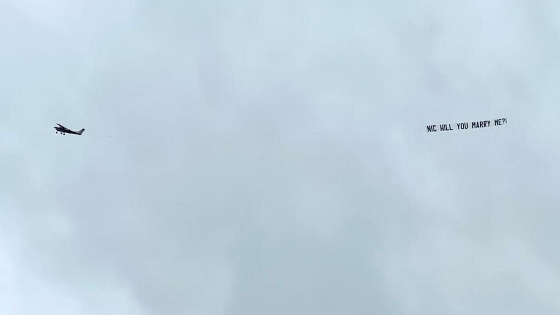 A small white plane carries a banner reading "Nic will you marry me?!" across a grey sky