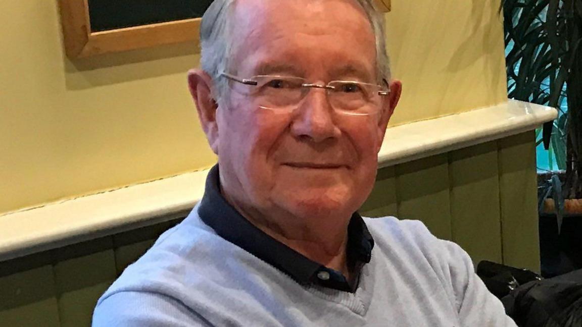 A man with grey hair and wearing glasses looking at the camera and smiling. He is wearing a light grey jumper with a yellow wall behind him. 