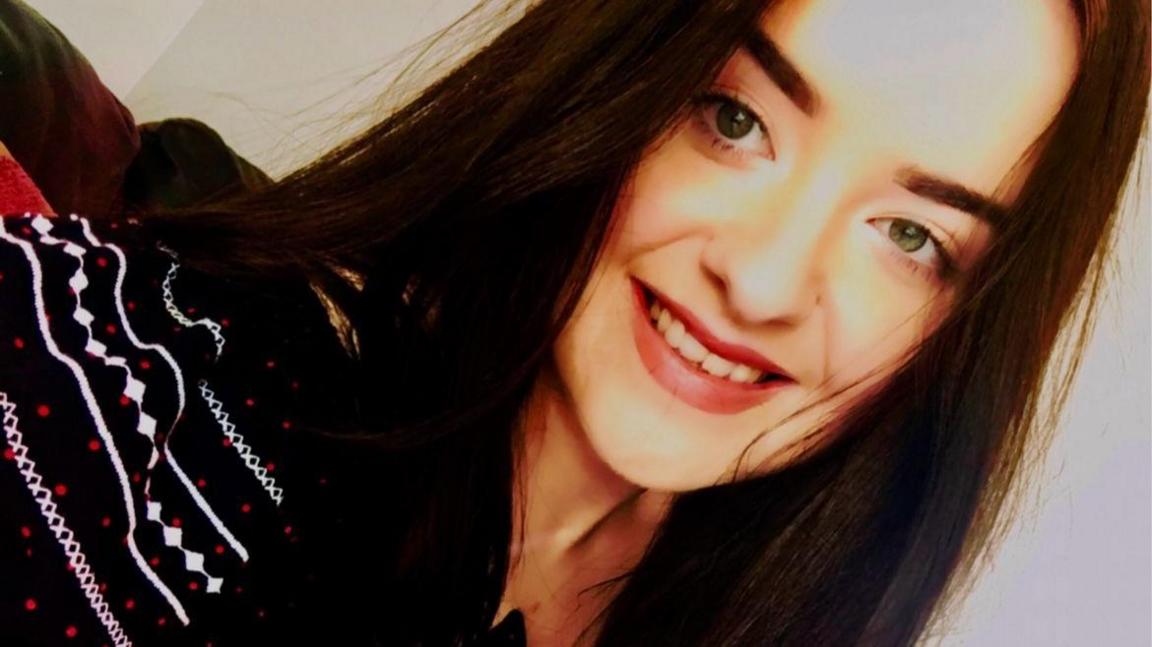 This is a selfie of Emily, who has long dark hair and is wearing a patterned, black top and smiling at the camera.