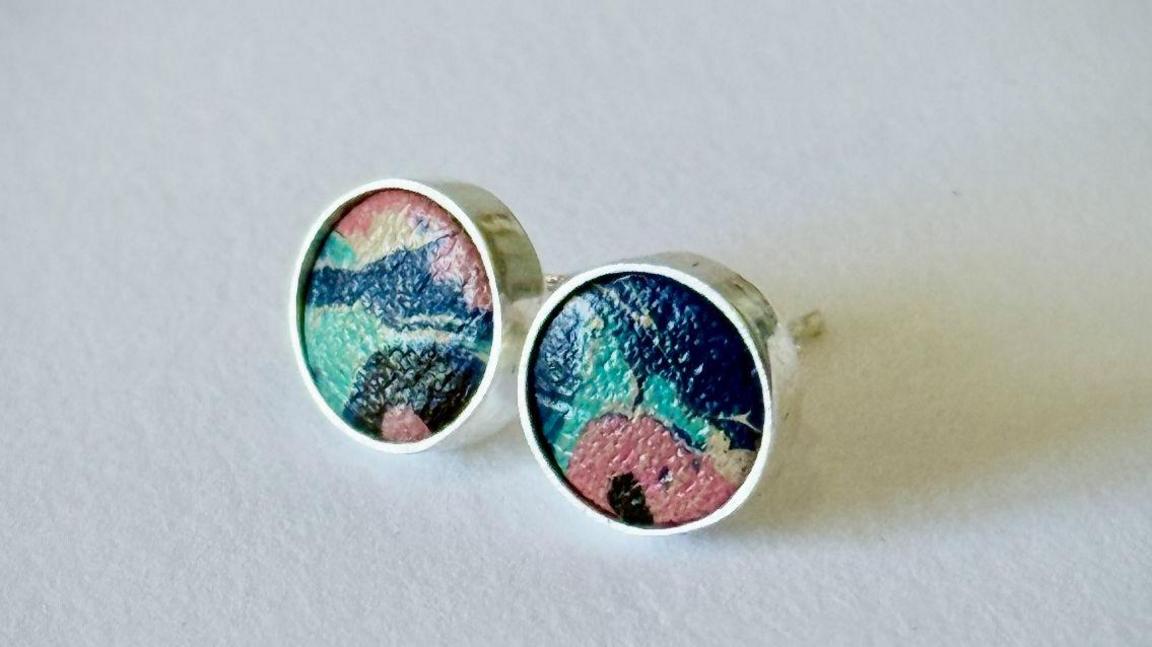 A circular stud earring contains a colourful piece of leather in the centre of it. The colours are green, blue and pink all marbled together.